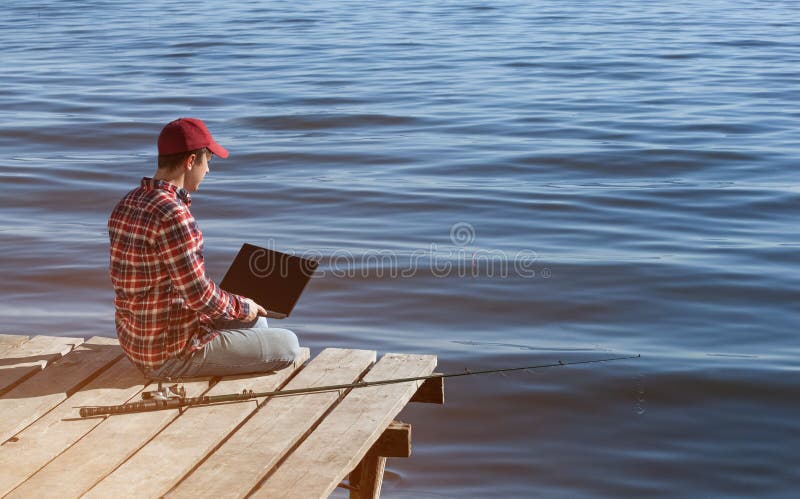 L'uomo del pescatore lavora ad un computer portatile, si siede su un pilastro di legno vicino al lago, accanto là è una canna da