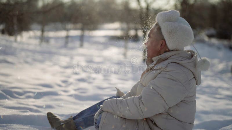 L'uomo felice è seduto a terra in inverno e ha gettato la neve per divertimento archivi video