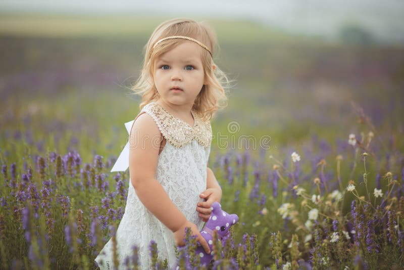 La bambina abbastanza sveglia sta portando il vestito bianco in un giacimento della lavanda che giudica un canestro pieno dei fio fotografia stock libera da diritti La bambina abbastanza sveglia sta portando il vestito bianco in un giacimento della lavanda che giudica un canestro pieno dei fio fotografia stock libera da diritti