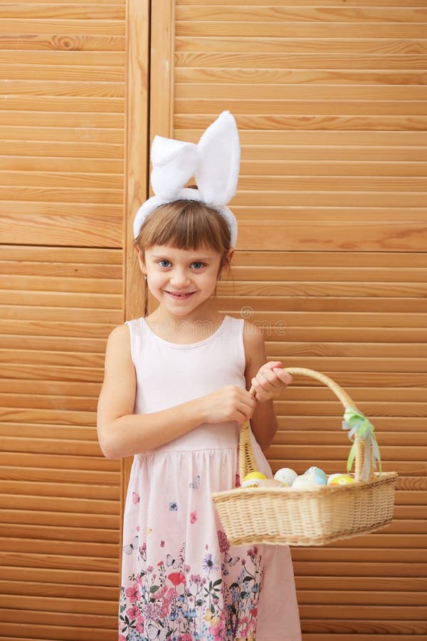 La bambina incantante nel vestito con le orecchie del coniglio bianche sulla sua testa tiene un canestro con le uova tinte sul fotografia stock La bambina incantante nel vestito con le orecchie del coniglio bianche sulla sua testa tiene un canestro con le uova tinte sul fotografia stock