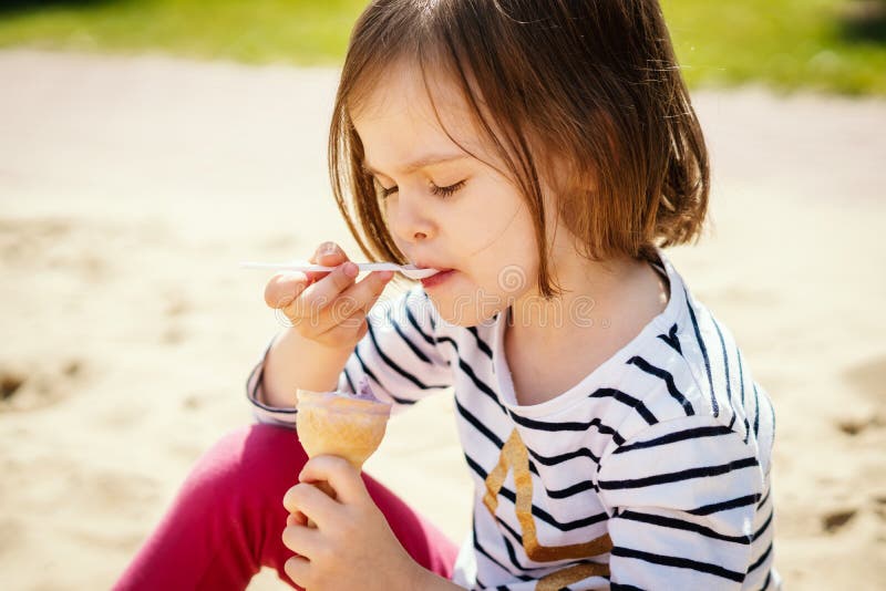 La bambina mangia il gelato che si siede sul campo da giuoco immagini stock libere da diritti La bambina mangia il gelato che si siede sul campo da giuoco immagini stock libere da diritti