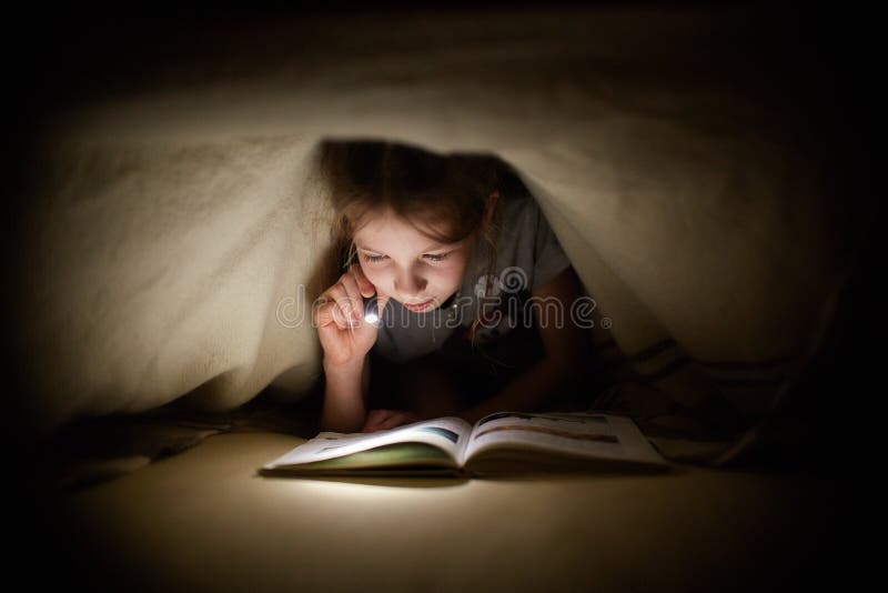 La bambina sta leggendo un libro sotto una coperta con una torcia elettrica in una stanza scura alla notte immagine stock libera da diritti La bambina sta leggendo un libro sotto una coperta con una torcia elettrica in una stanza scura alla notte immagine stock libera da diritti