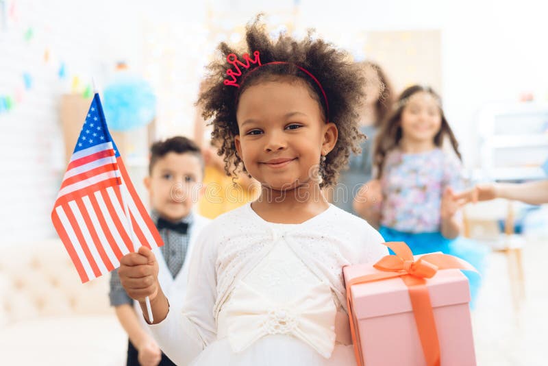 La bambina sveglia tiene un regalo e una bandiera di U.S.A. alla celebrazione del suo compleanno fotografia stock La bambina sveglia tiene un regalo e una bandiera di U.S.A. alla celebrazione del suo compleanno fotografia stock