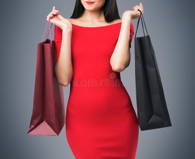 La bella donna in un vestito rosso sta tenendo i sacchetti della spesa operati Fondo grigio immagini stock La bella donna in un vestito rosso sta tenendo i sacchetti della spesa operati Fondo grigio immagini stock