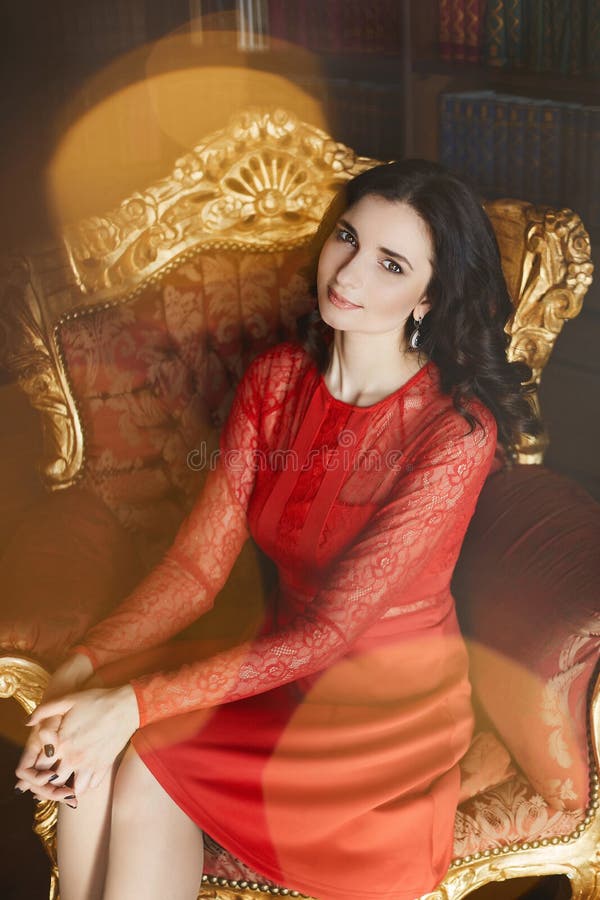 La bella e ragazza di modello castana affascinante con capelli ricci in vestito alla moda rosso si siede sulla poltrona d'annata immagine stock La bella e ragazza di modello castana affascinante con capelli ricci in vestito alla moda rosso si siede sulla poltrona d'annata immagine stock