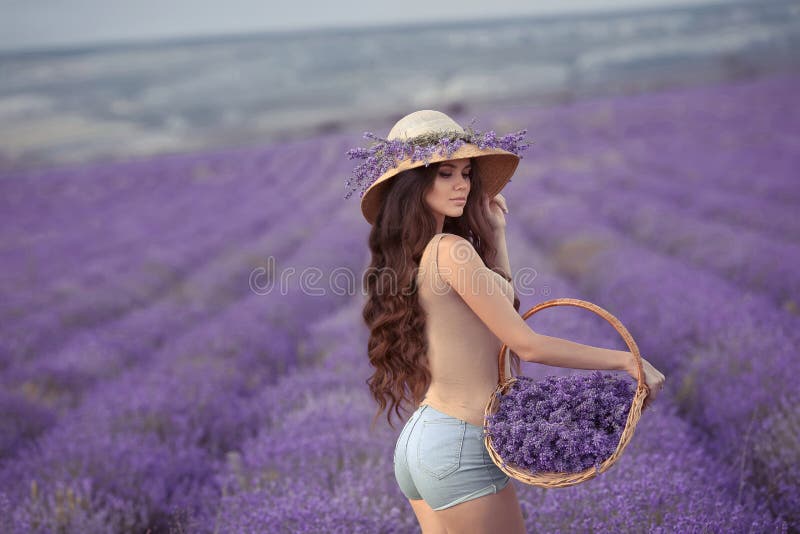 La bella giovane donna con nel cappello di vimini che posa nella porpora laven fotografia stock La bella giovane donna con nel cappello di vimini che posa nella porpora laven fotografia stock