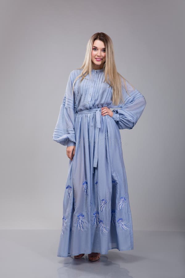 La bella giovane donna dresseed in vestito ricamato blu lungo sul fondo grigio dello studio immagine stock La bella giovane donna dresseed in vestito ricamato blu lungo sul fondo grigio dello studio immagine stock