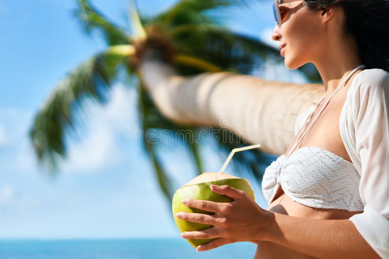 La bella giovane donna gode della bevanda della noce di cocco e si rilassa sulla spiaggia tropicale con la palma