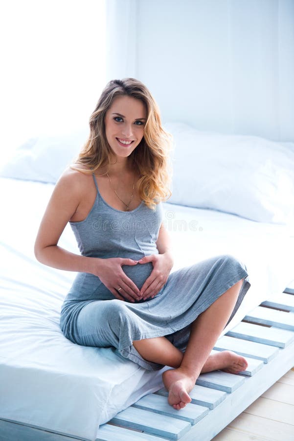 La bella giovane donna incinta si siede su un letto in una stanza e fa un cuore sulla sua pancia Luce solare luminosa di luce del fotografia stock La bella giovane donna incinta si siede su un letto in una stanza e fa un cuore sulla sua pancia Luce solare luminosa di luce del fotografia stock