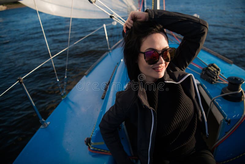 La bella giovane donna si siede sulla ciotola dell'yacht e posa Indossa i sunglusses con la mano ed il sorriso Il modello sta nav fotografia stock libera da diritti La bella giovane donna si siede sulla ciotola dell'yacht e posa Indossa i sunglusses con la mano ed il sorriso Il modello sta nav fotografia stock libera da diritti