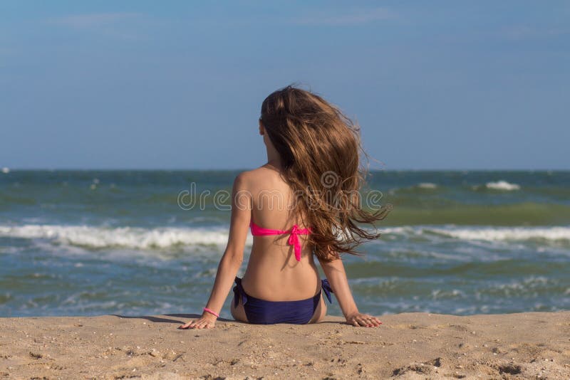La bella giovane donna in un costume da bagno con capelli lunghi si siede sulla spiaggia vicino al mare immagine stock La bella giovane donna in un costume da bagno con capelli lunghi si siede sulla spiaggia vicino al mare immagine stock
