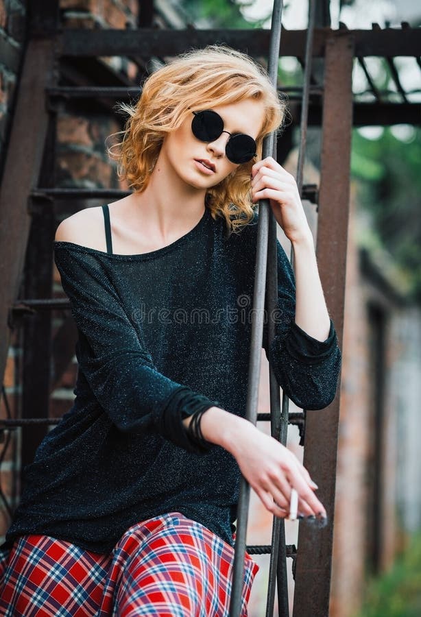 La bella giovane ragazza della roccia di lerciume che indossa la gonna a quadretti, il maglione ed i vetri si siede sulle scala e immagini stock La bella giovane ragazza della roccia di lerciume che indossa la gonna a quadretti, il maglione ed i vetri si siede sulle scala e immagini stock
