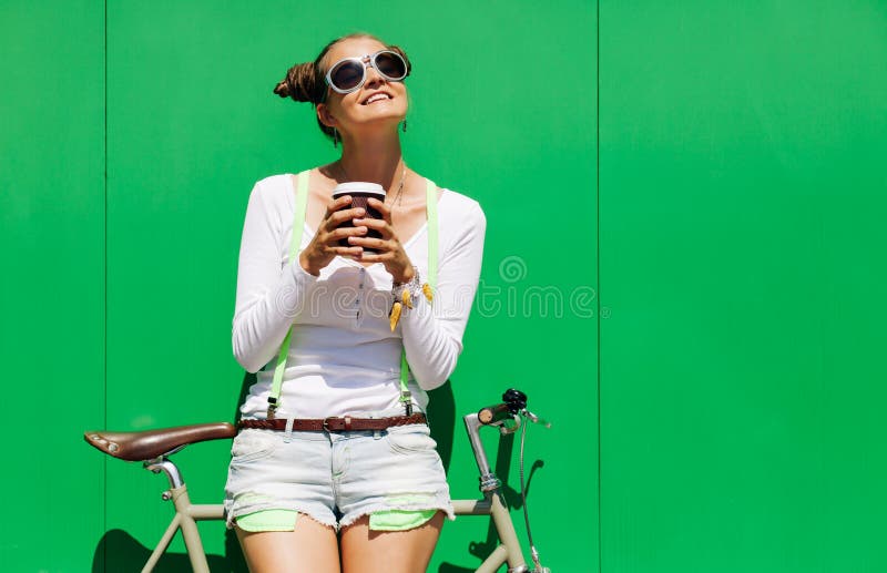 La bella giovane ragazza graziosa alla moda in breve e maglietta ed i supporti freschi degli occhiali da sole con la bicicletta r fotografia stock La bella giovane ragazza graziosa alla moda in breve e maglietta ed i supporti freschi degli occhiali da sole con la bicicletta r fotografia stock