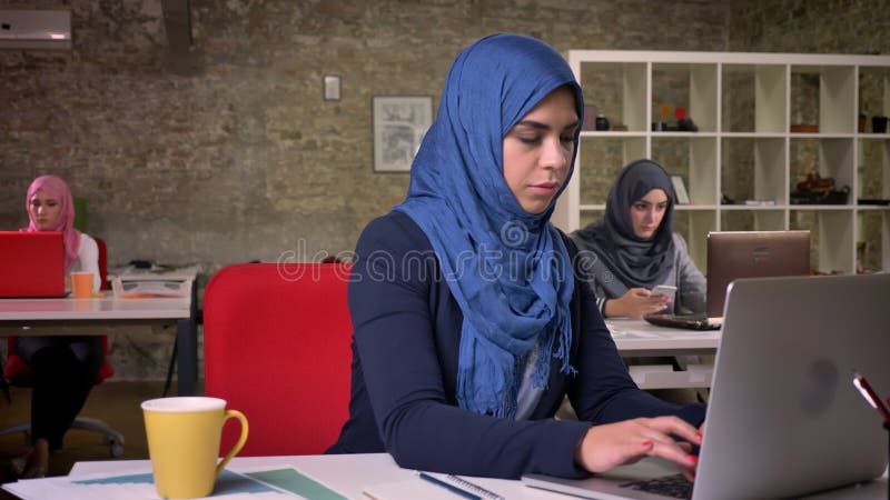 La bella ragazza araba lavorante astuta nel hijab blu scuro sta sedendosi a da tavolino e sta scrivendo sul suo computer, circond