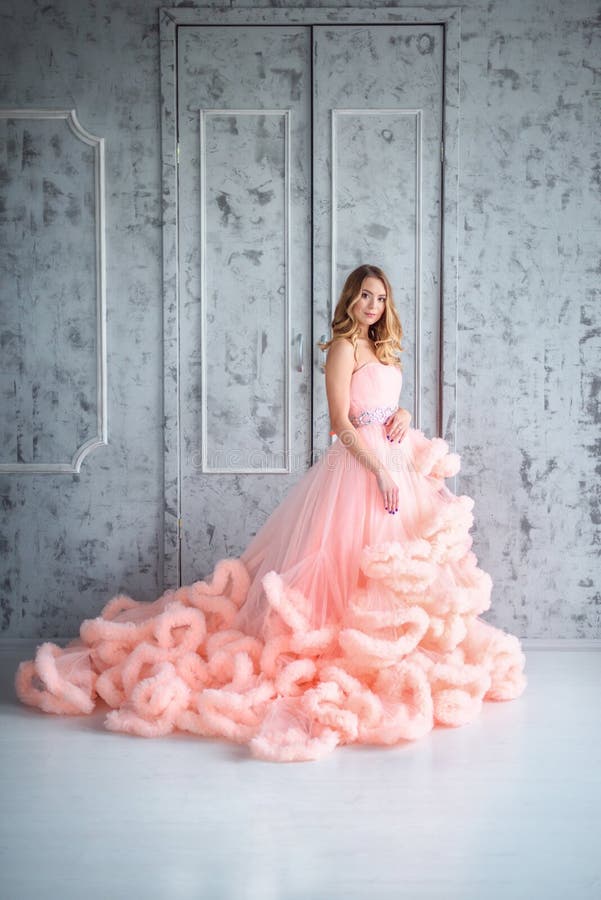 La bella ragazza con capelli ondulati lunghi che tengono un mazzo dei fiori in nuvola rosa del vestito- è nell'interno immagini stock La bella ragazza con capelli ondulati lunghi che tengono un mazzo dei fiori in nuvola rosa del vestito- è nell'interno immagini stock