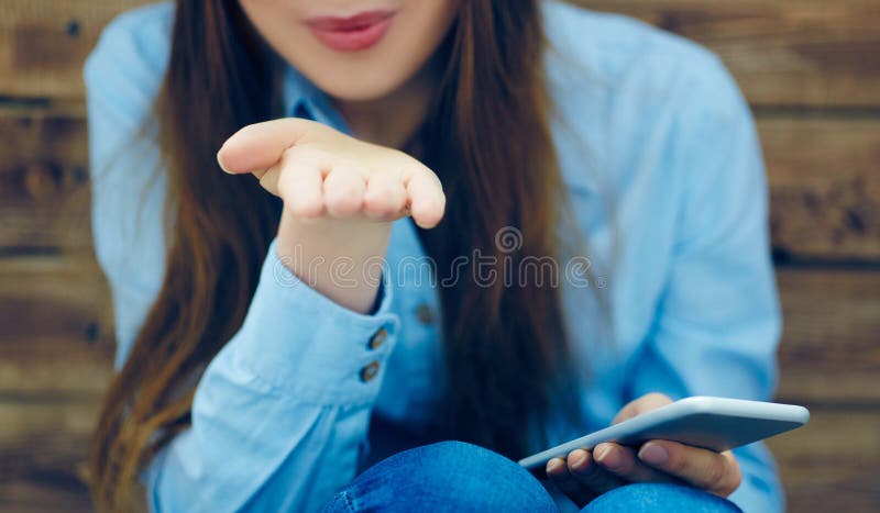 La bella ragazza con lo Smart Phone nelle manifestazioni delle mani ventila il gesto di bacio che si siede sui precedenti della p immagini stock libere da diritti La bella ragazza con lo Smart Phone nelle manifestazioni delle mani ventila il gesto di bacio che si siede sui precedenti della p immagini stock libere da diritti