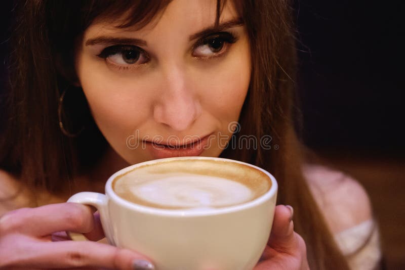 La bella ragazza dai capelli lunghi in un maglione bianco sta stando in una caffetteria ad una tavola di legno con una tazza, rit fotografia stock La bella ragazza dai capelli lunghi in un maglione bianco sta stando in una caffetteria ad una tavola di legno con una tazza, rit fotografia stock