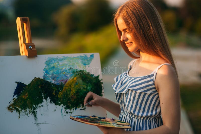 La bella ragazza disegna un'immagine nel parco facendo uso di una tavolozza con le pitture e una spatola Cavalletto e tela con un immagini stock La bella ragazza disegna un'immagine nel parco facendo uso di una tavolozza con le pitture e una spatola Cavalletto e tela con un immagini stock