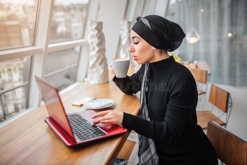 La belle et bien habillée jeune femme Arabe boit le café et des travaux sur l'ordinateur portable Elle s'assied à l'intérieur de image stock La belle et bien habillée jeune femme Arabe boit le café et des travaux sur l'ordinateur portable Elle s'assied à l'intérieur de image stock