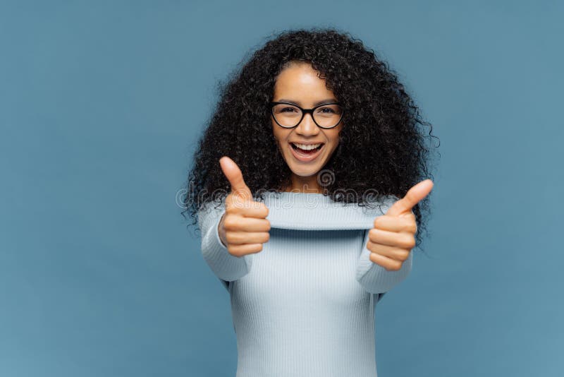 La belle femme heureuse avec les cheveux touffus d'Afro renonce au pouce, approuve l'idée gentille, utilise le chandail à la mode photo libre de droits La belle femme heureuse avec les cheveux touffus d'Afro renonce au pouce, approuve l'idée gentille, utilise le chandail à la mode photo libre de droits