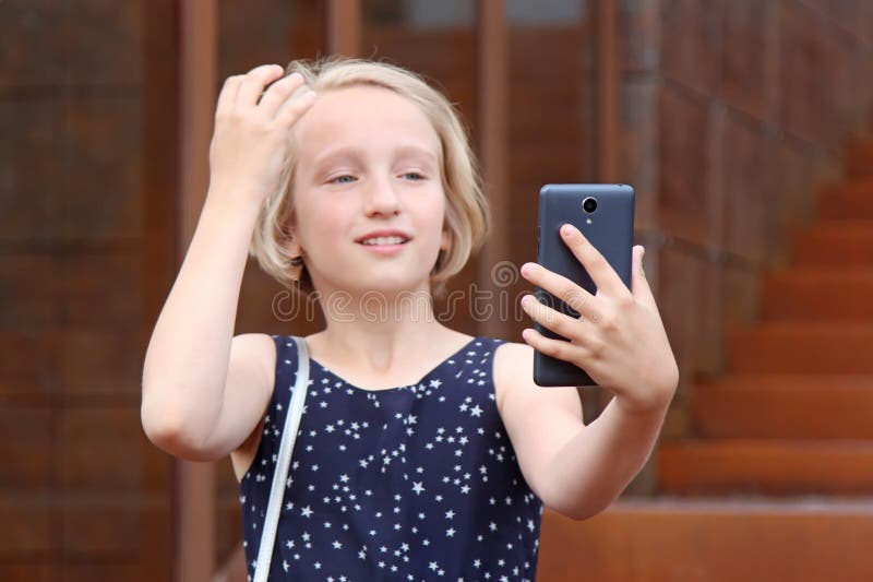 La belle fille de la préadolescence blonde à l'aide d'un téléphone, fait à la photo un appareil-photo avant, prenant un autoportr photographie stock libre de droits