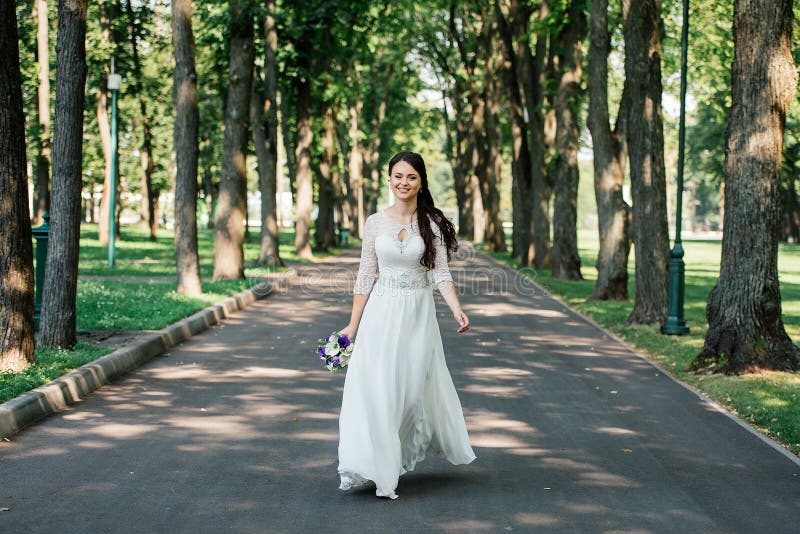 La belle jeune jeune mariée de sourire de brune dans la robe de wedd avec le bouquet des fleurs dans des mains entre en parc deho images stock La belle jeune jeune mariée de sourire de brune dans la robe de wedd avec le bouquet des fleurs dans des mains entre en parc deho images stock