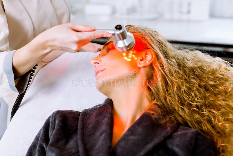 Beautician doing red led light therapy to blond women in beauty salon, facial photo therapy for skin pore cleansing. Anti-aging treatments and photo rejuvenation procedure, close up. Beautician doing red led light therapy to blond women in beauty salon, facial photo therapy for skin pore cleansing. Anti-aging treatments and photo rejuvenation procedure, close up.