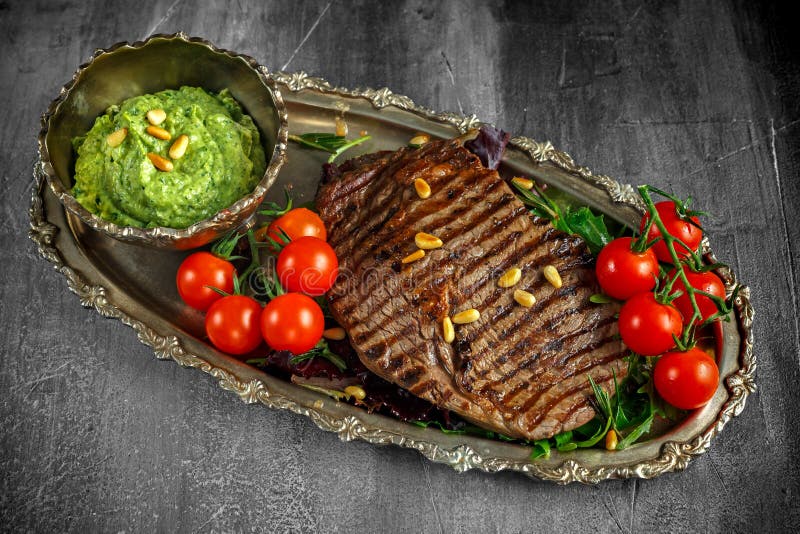 La bistecca arrostita di Ribeye è servito sul cuscino dell'insalata con il pesto, i pinoli ed i pomodori ciliegia su un piatto di fotografie stock libere da diritti La bistecca arrostita di Ribeye è servito sul cuscino dell'insalata con il pesto, i pinoli ed i pomodori ciliegia su un piatto di fotografie stock libere da diritti