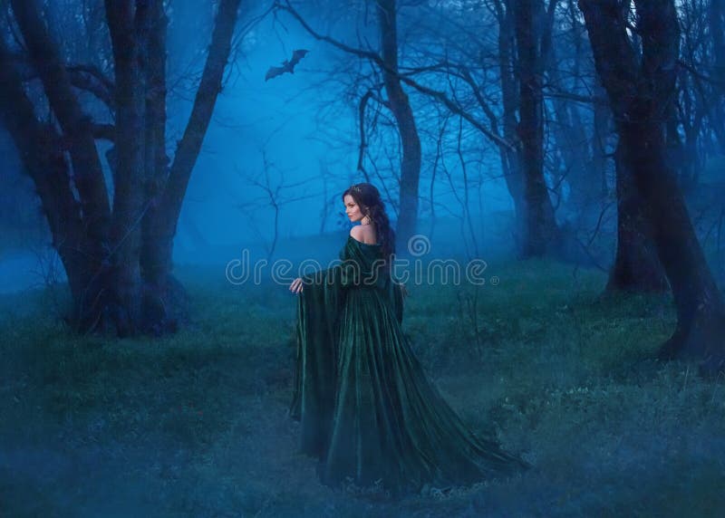 La bruja atractiva en un traje azul del terciopelo con un tren largo camina a través del bosque de la noche en busca de la víctim