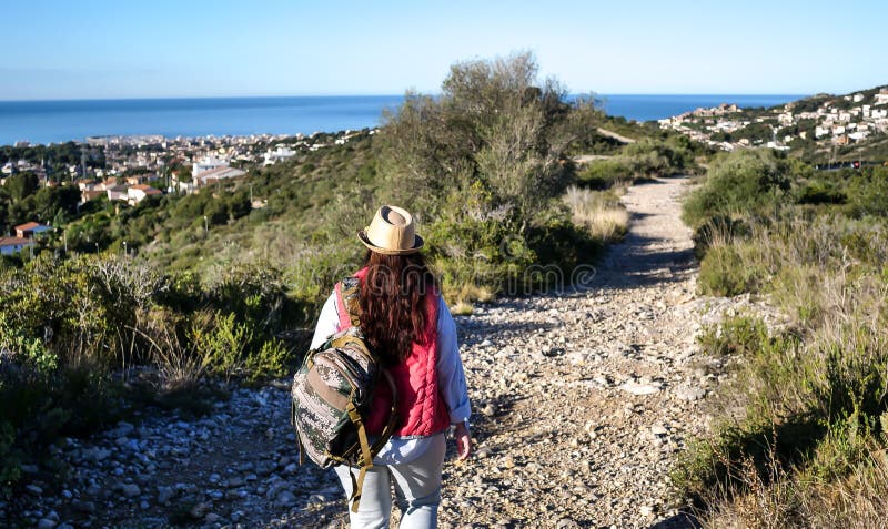 La brune de touristes dans un chapeau et un sac à dos va le long du chemin sur une colline à la ville par la mer photos stock La brune de touristes dans un chapeau et un sac à dos va le long du chemin sur une colline à la ville par la mer photos stock