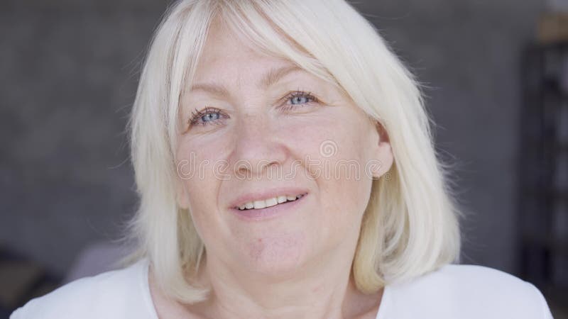 La cara de cerca de una hermosa mujer madura mirando la cámara. La mujer con los ojos azules y el pelo rubio en casa. Cámara almacen de metraje de vídeo