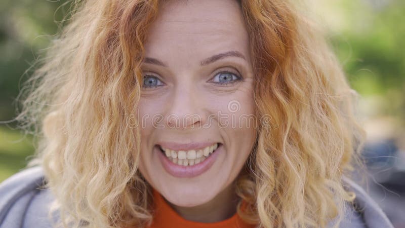 La cara de una linda mujer madura de cabello rojo con el pelo rizado sonriendo alegremente mirando la cámara en el parque almacen de video