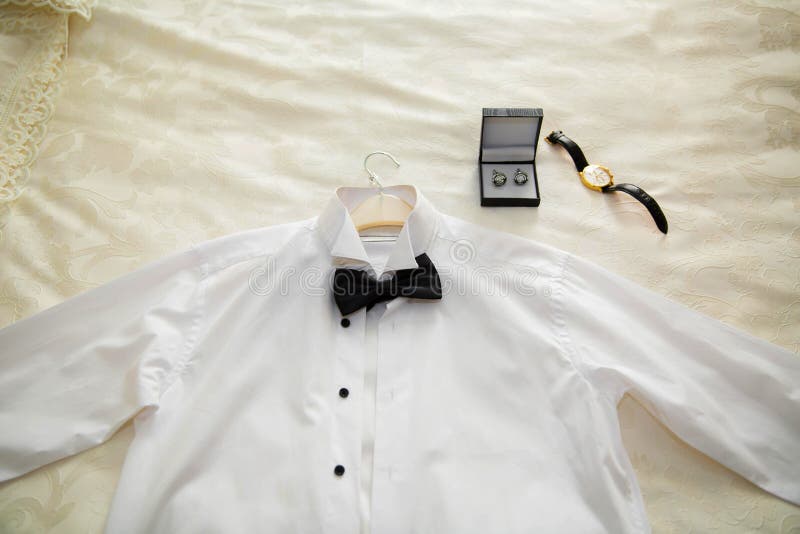 La chemise blanche, l'anneau, et la montre du bébé s'habillant sur le lit Chemises classiques du ` s d'hommes sur le lit photographie stock libre de droits La chemise blanche, l'anneau, et la montre du bébé s'habillant sur le lit Chemises classiques du ` s d'hommes sur le lit photographie stock libre de droits