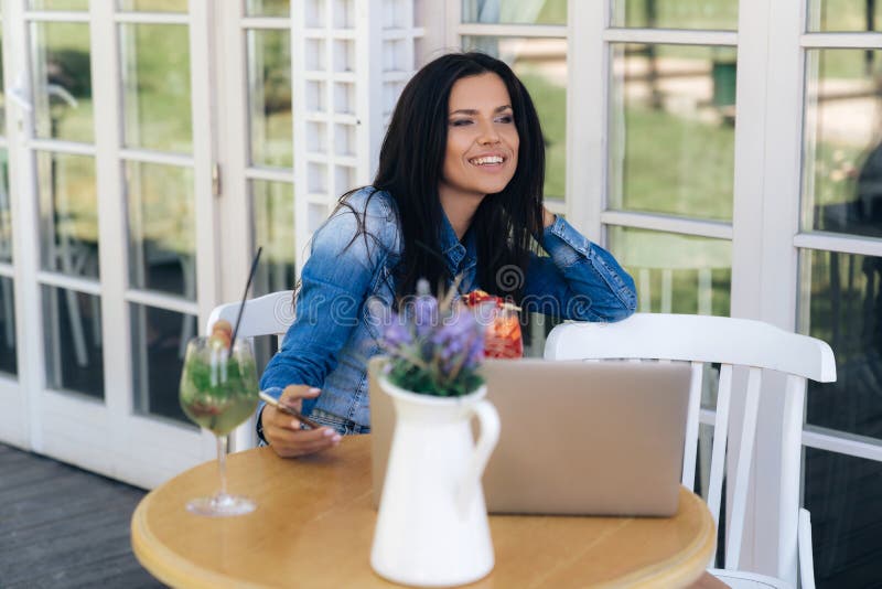 La chica joven atractiva sonriente se sienta en un café, risas, negociaciones alguien El modelo sostiene un smartphone, un ordena foto de archivo libre de regalías La chica joven atractiva sonriente se sienta en un café, risas, negociaciones alguien El modelo sostiene un smartphone, un ordena foto de archivo libre de regalías