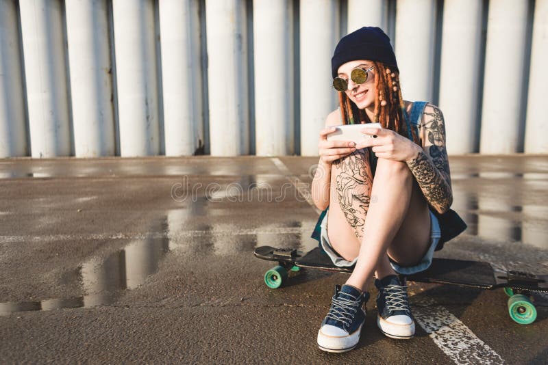 La chica joven con los tatuajes y los dreadlocks en un casquillo azul utiliza un smartphone contra la perspectiva de un muro de c foto de archivo libre de regalías La chica joven con los tatuajes y los dreadlocks en un casquillo azul utiliza un smartphone contra la perspectiva de un muro de c foto de archivo libre de regalías