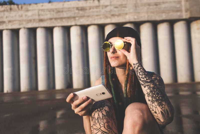 La chica joven con los tatuajes y los dreadlocks en un casquillo azul utiliza un smartphone contra la perspectiva de un muro de c foto de archivo La chica joven con los tatuajes y los dreadlocks en un casquillo azul utiliza un smartphone contra la perspectiva de un muro de c foto de archivo