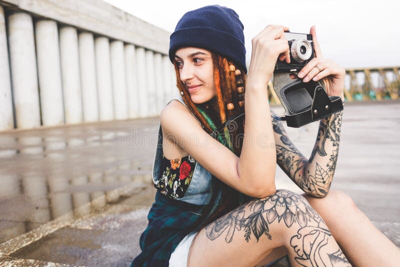 La chica joven con los tatuajes y los dreadlocks en un sombrero del azul fotografía una cámara del vintage en el fondo de un muro imagenes de archivo La chica joven con los tatuajes y los dreadlocks en un sombrero del azul fotografía una cámara del vintage en el fondo de un muro imagenes de archivo
