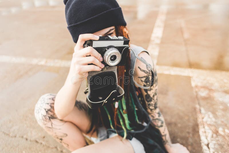 La chica joven con los tatuajes y los dreadlocks en un sombrero del azul fotografía una cámara del vintage en el fondo de un muro imágenes de archivo libres de regalías La chica joven con los tatuajes y los dreadlocks en un sombrero del azul fotografía una cámara del vintage en el fondo de un muro imágenes de archivo libres de regalías