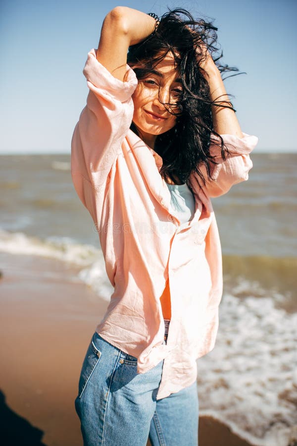 La chica joven hermosa con el pelo negro en una camisa y vaqueros se divierte en la playa del mar de Azov fotografía de archivo libre de regalías La chica joven hermosa con el pelo negro en una camisa y vaqueros se divierte en la playa del mar de Azov fotografía de archivo libre de regalías
