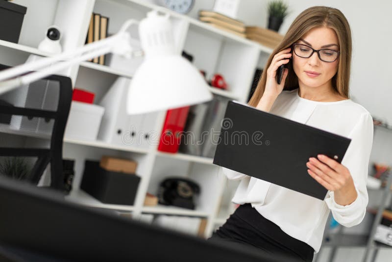 La chica joven se sentó en la tabla en la oficina, sosteniendo una tableta con las hojas en su mano y hablando en el teléfono fotografía de archivo La chica joven se sentó en la tabla en la oficina, sosteniendo una tableta con las hojas en su mano y hablando en el teléfono fotografía de archivo