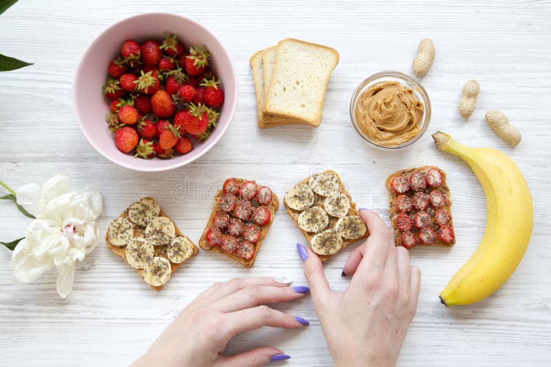 La chica joven toma la tostada del vegano con las frutas, semillas, mantequilla de cacahuete sobre el fondo de madera blanco, vis fotografía de archivo La chica joven toma la tostada del vegano con las frutas, semillas, mantequilla de cacahuete sobre el fondo de madera blanco, vis fotografía de archivo