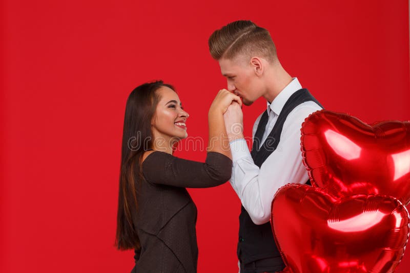 La coppia nell'amore, tipo bacia una mano del ` s della ragazza, accanto alle palle sotto forma di un cuore Fondo rosso immagine stock La coppia nell'amore, tipo bacia una mano del ` s della ragazza, accanto alle palle sotto forma di un cuore Fondo rosso immagine stock