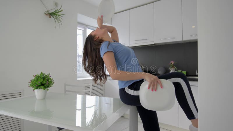 La danse femelle de femme au foyer drôle et chante avec des plats dans des bras tout en faisant cuire le repas à la cuisine à la