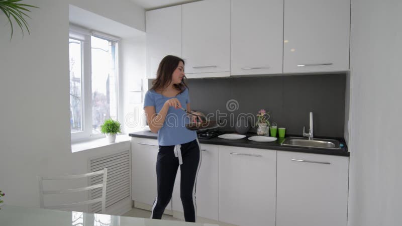 La danse gaie de femme de charge et chante avec la casserole et les plats dans des mains tout en faisant cuire le repas à la cuis