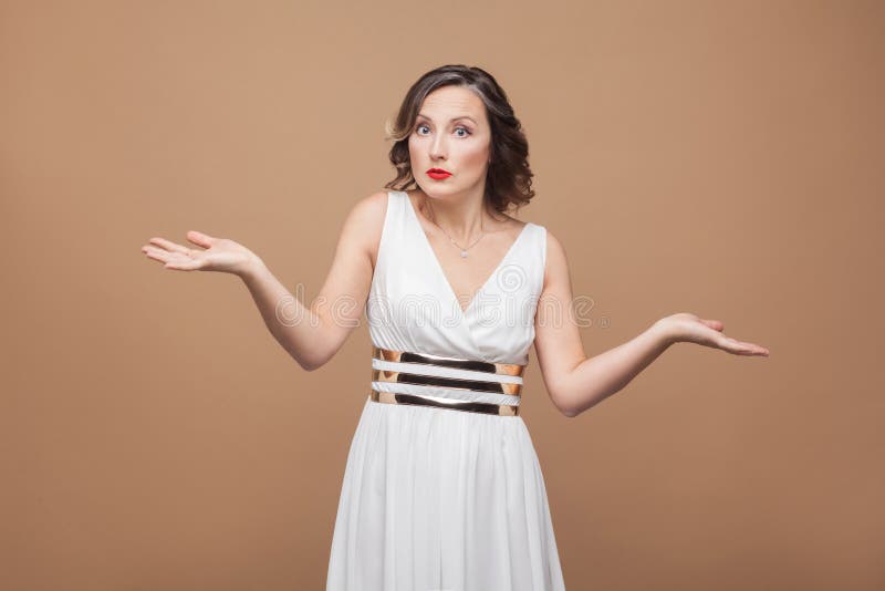 Puzzled adult woman say i don`t know. Emotional expressing woman in white dress, red lips and dark curly hairstyle. Studio shot, indoor, on beige or light brown background. Puzzled adult woman say i don`t know. Emotional expressing woman in white dress, red lips and dark curly hairstyle. Studio shot, indoor, on beige or light brown background