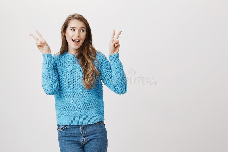 Cheerful woman with positive expression raising hands and showing victory or peace gestures, looking aside while singing, standing against gray background. Girl makes air quotes while being sarcastic. Cheerful woman with positive expression raising hands and showing victory or peace gestures, looking aside while singing, standing against gray background. Girl makes air quotes while being sarcastic.