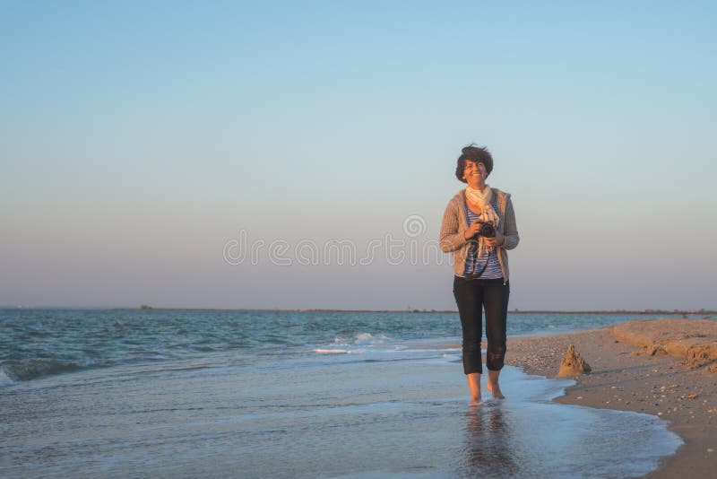 La donna allegra sta camminando lungo la spiaggia con la macchina fotografica fotografia stock La donna allegra sta camminando lungo la spiaggia con la macchina fotografica fotografia stock
