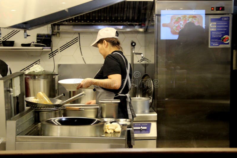 La donna asiatica prepara l'alimento dietro un divisore in vetro, corte di alimento al mercato centrale fotografia stock La donna asiatica prepara l'alimento dietro un divisore in vetro, corte di alimento al mercato centrale fotografia stock