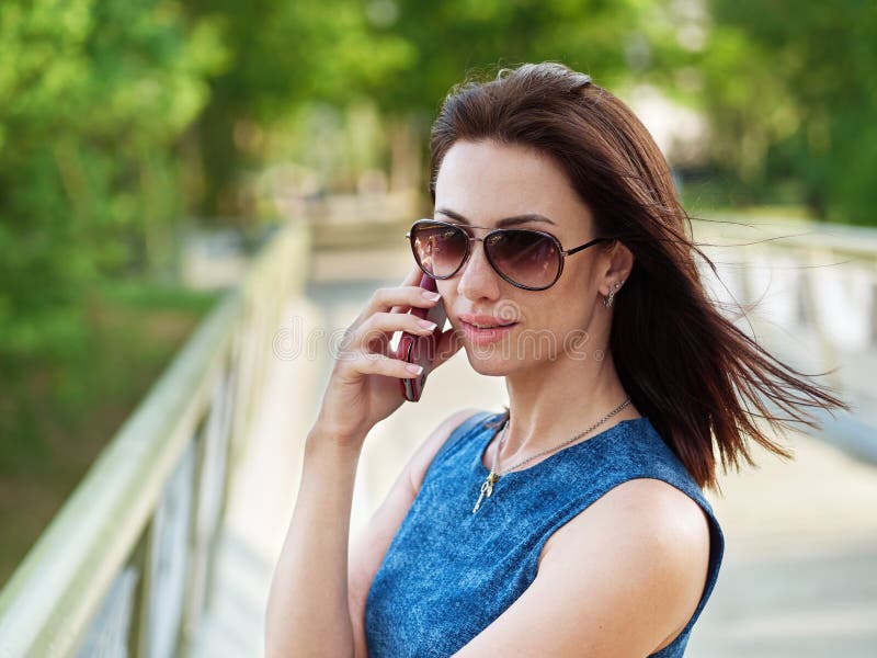 La donna castana attraente in occhiali da sole e vestito dalle blue jeans fa battersi la conversazione telefonica emozionale sul immagini stock La donna castana attraente in occhiali da sole e vestito dalle blue jeans fa battersi la conversazione telefonica emozionale sul immagini stock