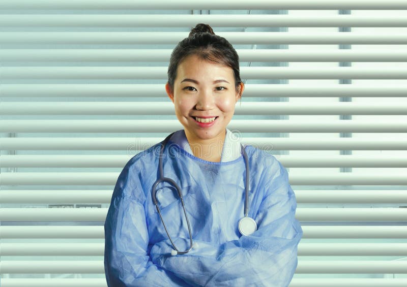 La donna coreana asiatica attraente di medico della medicina in blu sfrega la posa allegra sorridente corporativa alla finestra d fotografia stock libera da diritti La donna coreana asiatica attraente di medico della medicina in blu sfrega la posa allegra sorridente corporativa alla finestra d fotografia stock libera da diritti